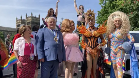 STUART PRANDLE/BBC Mayor Gary Hutchinson, wearing a blue suit and his golden chain of office, stood with three drag queens taking part in the march. They wear dramatic dresses. Notably, one wears a dress designed to look like orange flames, with a golden crown fashioned to look like a blooming flower.
