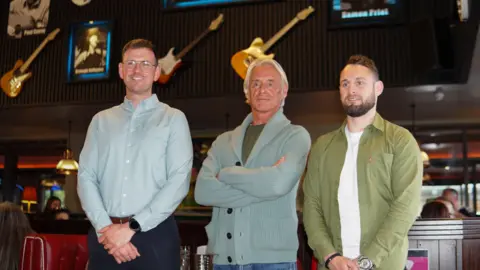 Nathan Edgar Three men stand in front of a wall on which guitars hang. The man on the right is tallest and wears a light blue shirt, the man in the middle has longish blond hair and is wearing a green cardigan, while the man on the right is weraing a green jacket and white t shirt