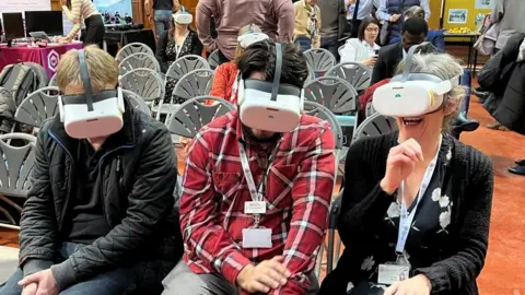 Ollie Conopo/BBC Three people wearing white VR headsets. The one on the right is laughing and appears to be raising her hand to touch something that is not there.