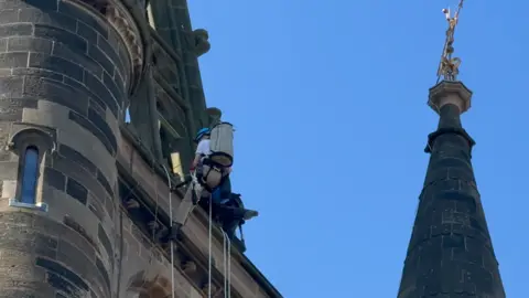 Clarke Elsby A man wearing a harness scales an old Gothic building tower