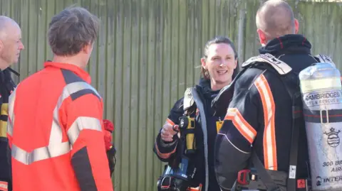 Cambridgeshire Fire and Rescue Service Ms Bourne is talking to three male firefighters. They are facing away from the camera - towards her. She is wearing her firefighting uniform and carrying equipment.