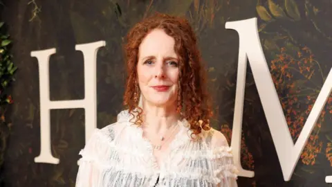Maggie O'Farrell has curly red hair which is half up. She is wearing a sheer white blouse with a black tank top underneath. Behind her can be seen a large white H and M on a green forest background. 