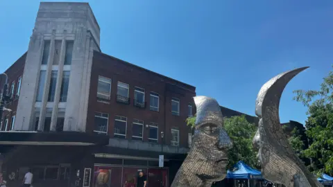 A public square with a large silver sculpture of two faces staring at each other. Behind the sculpture are brick buildings, trees, market stalls with blue gazebos, and a few people walking. The scene is illuminated by bright sunshine beneath a clear blue sky.