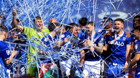 PA Media A group of football layers wearing blue and white football kits standing on a football pitch celebrating. There are white and blue streamers falling down from the sky.