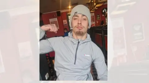 A young man, wearing a grey zipped hoodie and grey woolly hat is sitting at a table in a pub, flexing his right arm to show his muscle. 