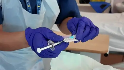 Medical staff wearing purple nitrate gloves holds a syringe inside a hospital setting.