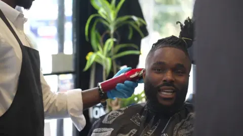 BBC News A staff member at Jul's barber shop in south London looks at a customer while testing his blood pressure with an out-of-shot machine that has a cuff