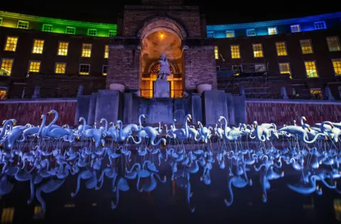 Hundreds of illuminated model flamingos are visible in front of City Hall in Bristol. They are in the water in front of the building's main entrance