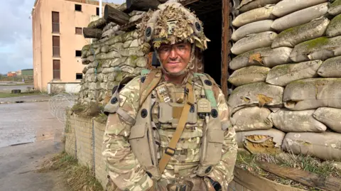 Major Lance Morris in army wear with flak jackets and a satchel. He is wearing a helmet covered in fake leaves and is standing outside a building which has fake leaves and wooden slats 