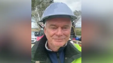 Contributed James Forsyth smiles at the camera. He wears a blue hard helmet on his head as well as a hi-vis jacket over a green coat. A flat bed truck can be seen parked behind him.