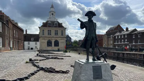 Ship chains lay along a quayside. Buildings are across the water and a statue of a man wearing a tricorn three-cornered hat stands on a grey, stone plinth. 