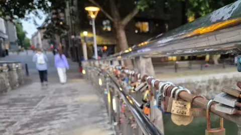 LDRS/Alex Seabrook A long line of padlocks stretches along the handrail of Pero's Bridge. Two people are walking along the bridge in the distance. Street lights are lit.