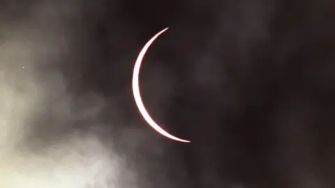 The moon has moved across the sun leaving a thin band of the sun visible which has taken on a pink hue. The sky is cloudy.