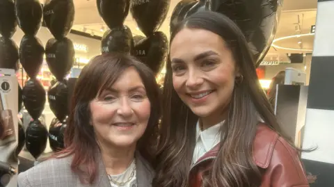 Standing side by side, Kayleigh has shoulder length brown hair and is wearing a grey blazer. Angela has long brown hair and is wearing a red patent jacket.