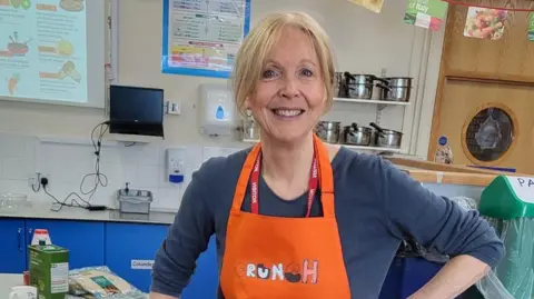 Kate smiles at the camera in a school kitchen classroom. She has blonde hair, tied up, and wears an orange apron with the CRuNCH logo. 