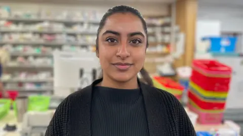 BBC/Gemma Dillon Trainee pharmacist Roshni Landa stands in front of drug supplies and trays at a pharmacy in Leeds