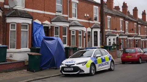 A police car parked at the side of a residential street. One of the terraced homes has blue material across the entrance.