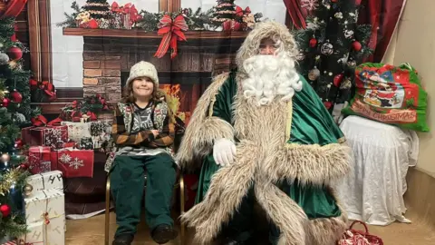 Karl Hasketh A child wearing a knitted hat and jumper sits beside a man dressed as St Nicholas, who is dressed in a green robe with a long white beard. They are indoors in front of a festive backdrop. 