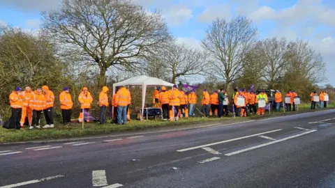 Paul Moseley/BBC About 40 Unison members dressed in hi-vis orange jackets standing by the A140 at Aylsham