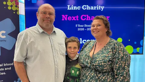 A family of three - a middle aged man and woman and their eight-year-old son, smile as they stand in front of promotional boards and screens for Linc, the blood cancer charity. The man is wearing a pale blue short-sleeved shirt and is bald, and the woman is wearing a dark green v-neck, long-sleeved dress with light orange coloured flowers on it. Their son, stood between them, is wearing a Minecraft Creeper T-Shirt. They are all smiling.