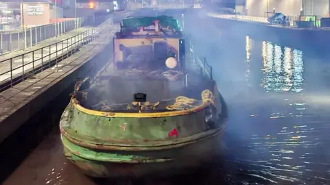 Humber Rescue Shot of the barge moored up after being freed. It is dark and there is a mist across the water, with buildings in the background.