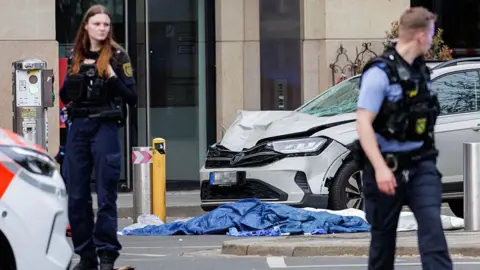 AFP via Getty Images Two out of focus police officers standing in front of a white car with a broken window and bent hood with blue and white tarps on the ground.