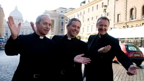 Getty Images Three priests - Fr Eugene O'Hagan, his brother Fr Martin O'Hagan and Fr David Delargy wearing priest vestments and collars. They are stood with their arms out in the middle of the street as they pose for the camera.