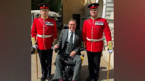 Handout A man, wearing a grey suit, is sitting in a wheelchair. There are two guards, in red coats, standing beside him.