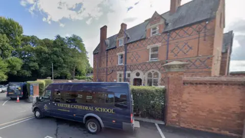 An street view of outside of Exeter Cathedral School. There is a blue bus outside of the building with the words 'Exeter Cathedral School'. The building is brown with blue details.