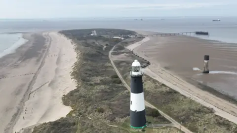 A black and white lighthouse stands in the middle of a spit - or peninsula, with the North Sea on one side and the Humber Estuary on the other. Vegetation and a single track road cover the central parts of the land. 