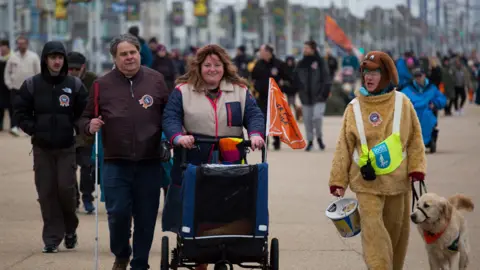 Sarah Summerton Guide dog user Mark Fielding, with guide dog trainer Gemma Fairhurst, push Ian in a pram along Blackpool Promenade. A charity fundraiser, dressed in a dog costume, holds a charity collection bucket as she walks alongside them.