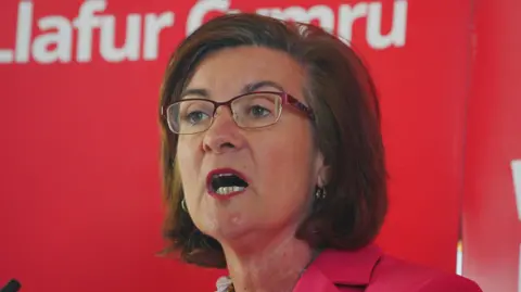 PA Media Eluned Morgan wearing glasses and a pink jacket giving a speech in front of a Welsh Labour backdrop.