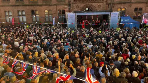 Crowds watching Si King and Lili Myers give a speech in Barrow. Union flag bunting can be scene and a stage.
