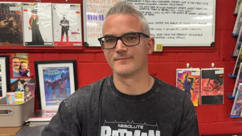 A man with white hair, glasses and black Batman top looking at the camera