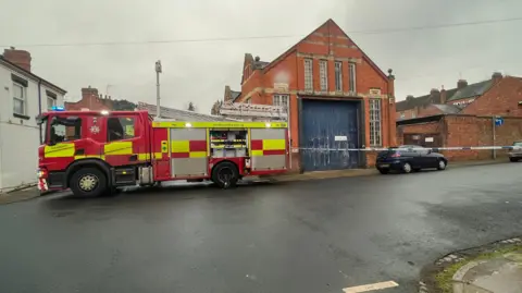 Kate Bradbrook/BBC A fire engine parked next to an old industrial-type building with large blue wooden doors. There is a cordon going across the street.