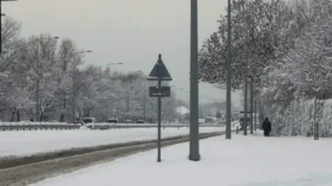 Keith Jones The A59 in 2010 is down to one lane with thick snow on pavements each side of the road. Cars are driving slowly in the distance while a lone figure in black walks along the former pavement.
