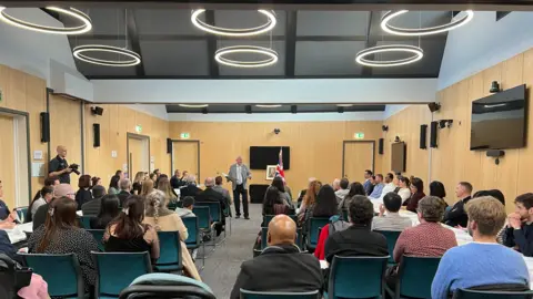 A man dressed in a grey and black suit addresses a room full of people with a picture of the King in the background