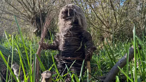 A small wicker man with long hair on a boat in amongst the reeds at a nature reserve.