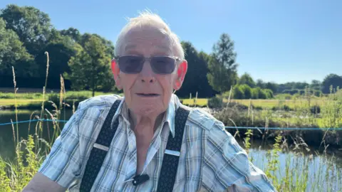 An elderly man wearing a checkered shirt and braces. He has white hair and is wearing sunglasses. He is sat looking into the camera, with a pond, fields and trees behind him