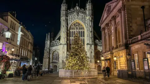 Getty Images A large illuminated Christmas tree is seen in front of Bath Abbey in the city centre. Small groups of people are looking at the tree. The photograph is taken at night with lights from local businesses glowing and the abbey also lit up