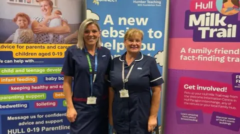 NHS Humber Two women in dark-blue nursing uniforms smiles as they stand in front of three colourful display boards containing information about breastfeeding. The woman on the left has blond, shoulder-length hair and wears a green and blue lanyard with identity card. The woman on the right has short strawberry-blond hair and wears a white lanyard. One of the boards offers "advice for partens and carers with children aged 0-19"; another advertises "Hull Milk Trail".