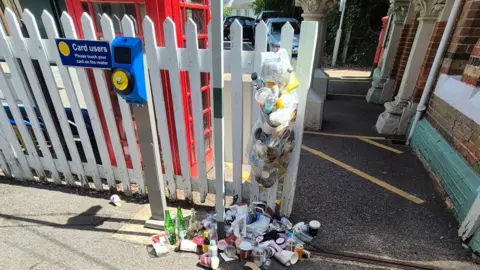 Overflowing rubbish hangs from a fence at Box Hill & Westhumble railway station on 5 July 2026. Plastic bottles, coffee cups, cans and packaging have spilled onto the ground beside a ticket validator and station entrance.