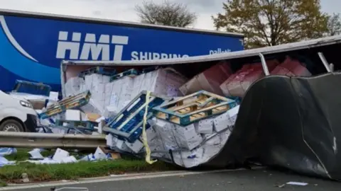 The scene of the crash on the M6 in Lancashire. A trailer straddles the central reservation, with pallets covered by tarpaulins. A blue lorry and the front of another vehicle are in the background.