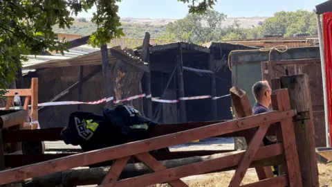Fire-damaged farm building cordoned off with safety tape, viewed behind a wooden gate. A firefighter's jacket rests on the gate, with a person standing nearby and scorched structures visible in the background.