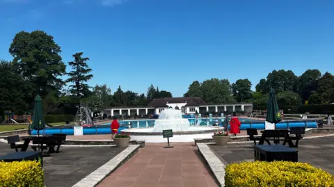 A paved path at a lido leads to a fountain, which sits in front of a big blue swimming pool. Behind the pool, there is a white poolhouse. The lido is set in a park, with trees surrounding it.