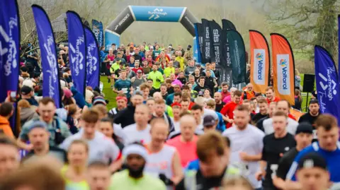A view of a field of runners starting the half marathon. There is a soft focus with the runners in the foreground blurred while those towards the back in sharp focus. The inflatable arch at the start is blue and black and there are advertising flags lining the sides of the route.