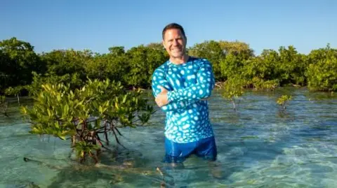Steve Backshall wearing a blue waterproof short and shorts standing thigh-high in crystal clear water