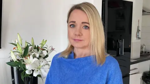 A woman with shoulder length blonde hair is wearing a blue shawl. She is looking into the camera and standing in a kitchen. Behind her is a vase of flowers and the kitchen looks modern and black and white.