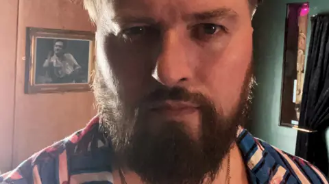 A close-up photo of a man with a beard who is wearing clothing of various colours. Curtains are visible on the far right, near to a green wall.