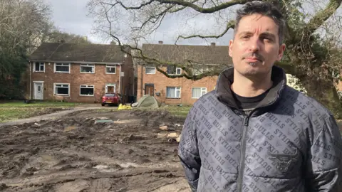 local resident Pedro Oliveira is wearing a black jacket and is standing in front of some muddy waterlogged land, in front of some beige-bricked houses.